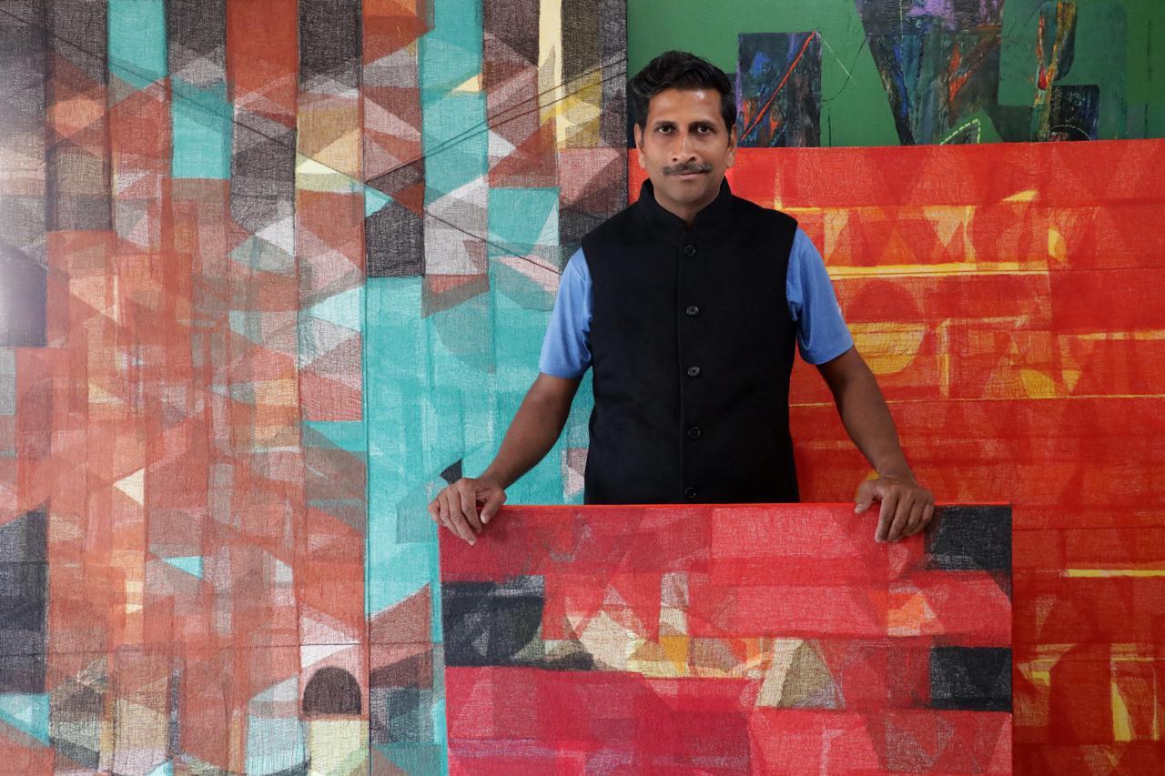 An Indian man with a mustache, identified as artist Vivek Nimbolkar, stands in his art studio. He is dressed in a blue polo shirt and a black Nehru vest. He holds a rectangular canvas featuring a red and orange geometric abstract painting. Behind him are several larger vertical canvases with intricate, multi-colored abstract designs consisting of overlapping triangles, rectangles, and textured earthy tones.